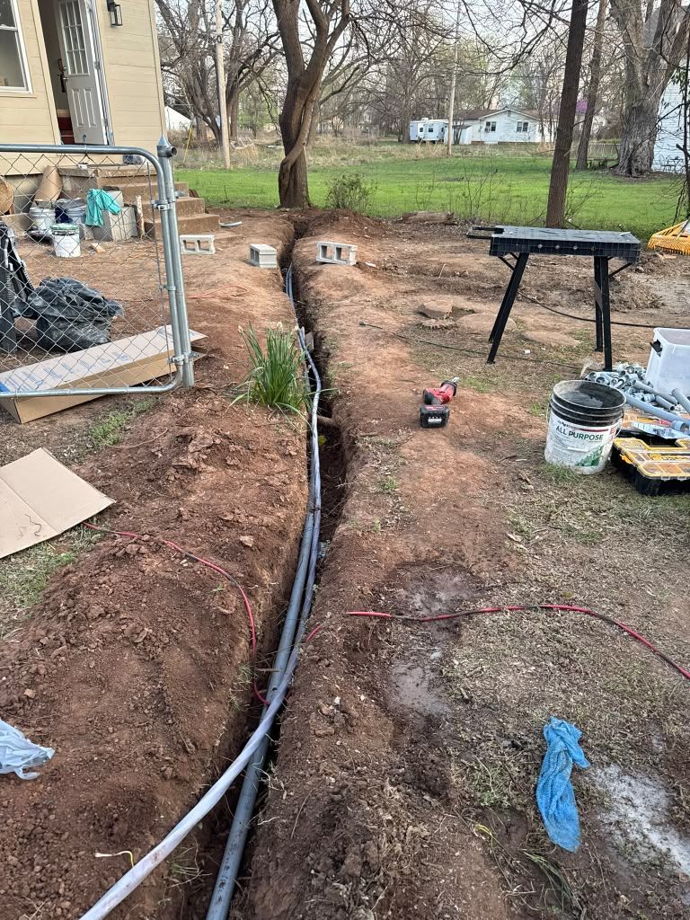Backyard trench with buried black pipe, red dirt, tools, and a table near a house and trees