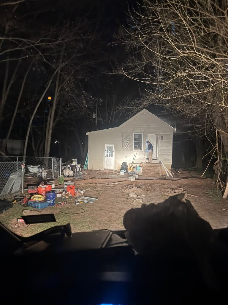 Small white house at night, lit by headlights, with scattered items and bare trees in the yard.