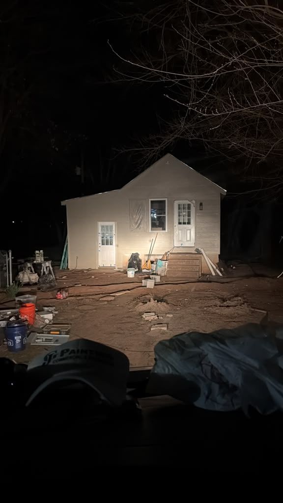 Nighttime construction scene with a lit house exterior, workers, and scattered building materials.