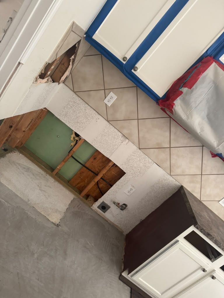 Tilted view of a damaged kitchen with exposed wall framing, tiled floor, and a white cabinet door open.