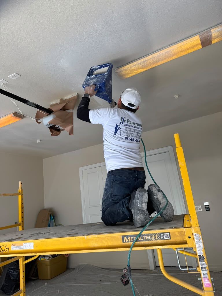 Worker kneeling on a scaffold, sanding a ceiling patch with a power sander in a room under renovation.
