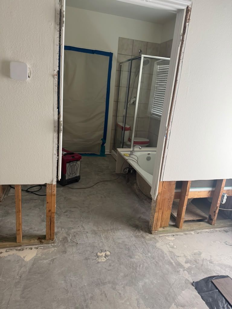 Unfinished bathroom doorway with exposed studs, gray floor, and taped-off shower or tub area.