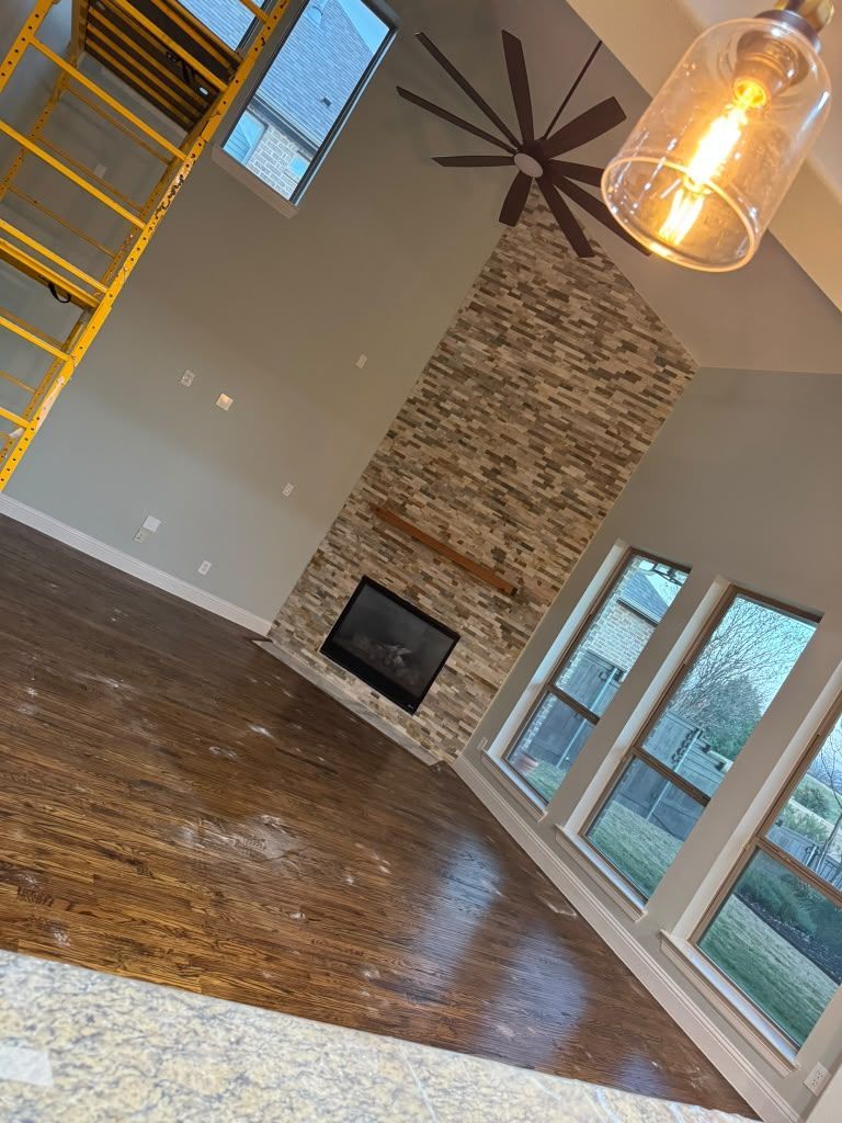Empty living room with hardwood floors, stone fireplace, ceiling fan, and large windows.