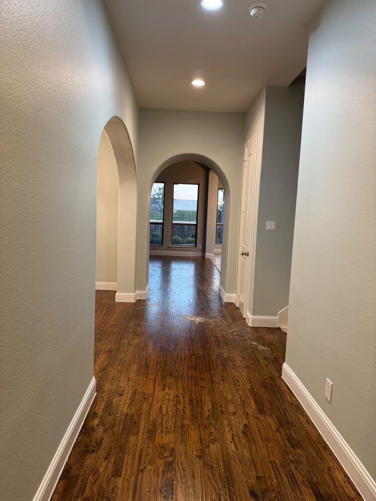 Empty hallway with hardwood floors, arched doorways, and recessed ceiling lights leading to a bright room.