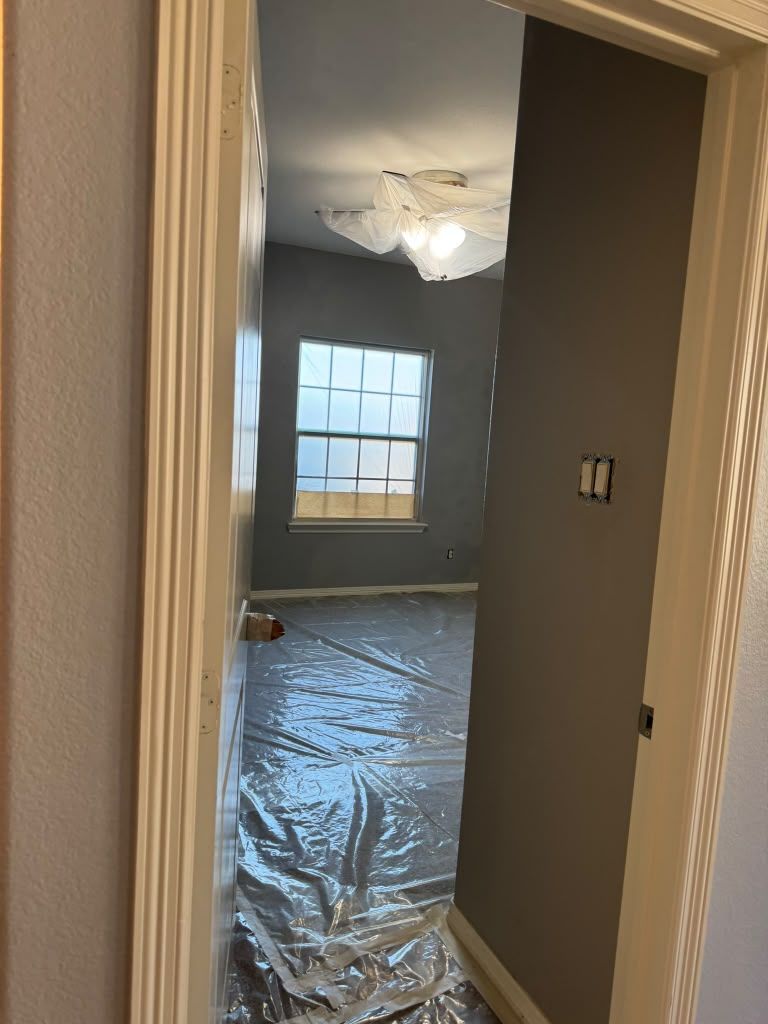 View through a doorway into a room under renovation, with plastic-covered floor, window, and damaged ceiling light.