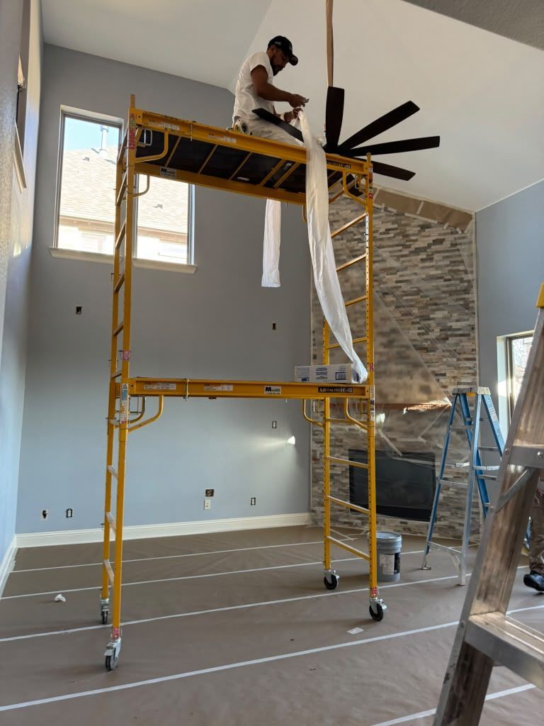 Worker on yellow scaffolding painting a room with high ceiling and fan.