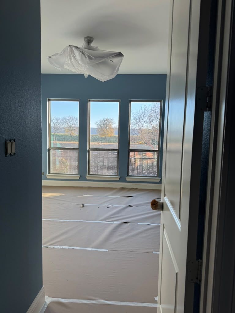 Empty blue room with large windows, covered floor, and a hanging light fixture brace