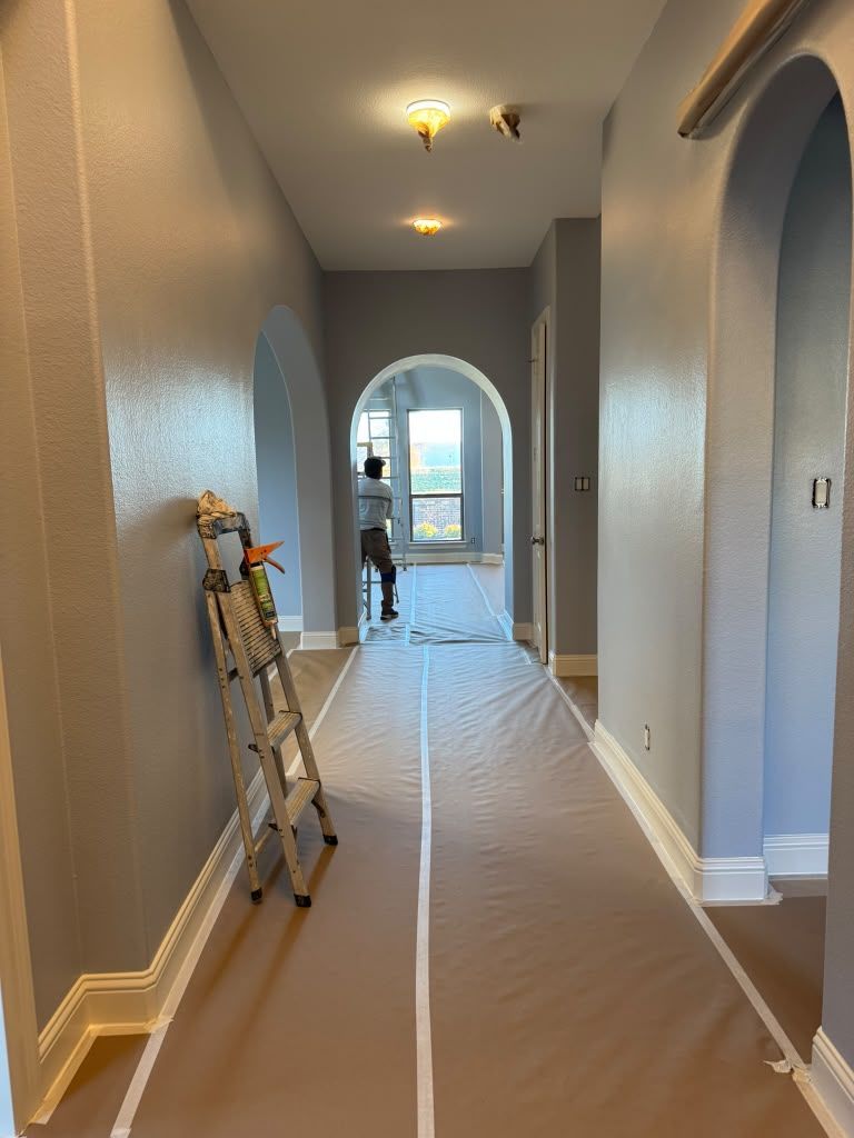 Hallway under renovation with plastic floor covering and a ladder against the left wall, lit by ceiling fixtures.