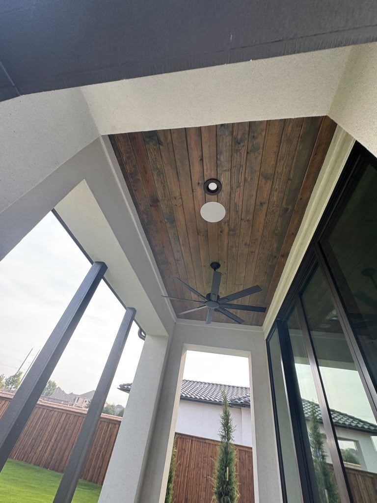Covered outdoor patio with wood-paneled ceiling, ceiling fan, recessed lights, and glass walls overlooking a yard