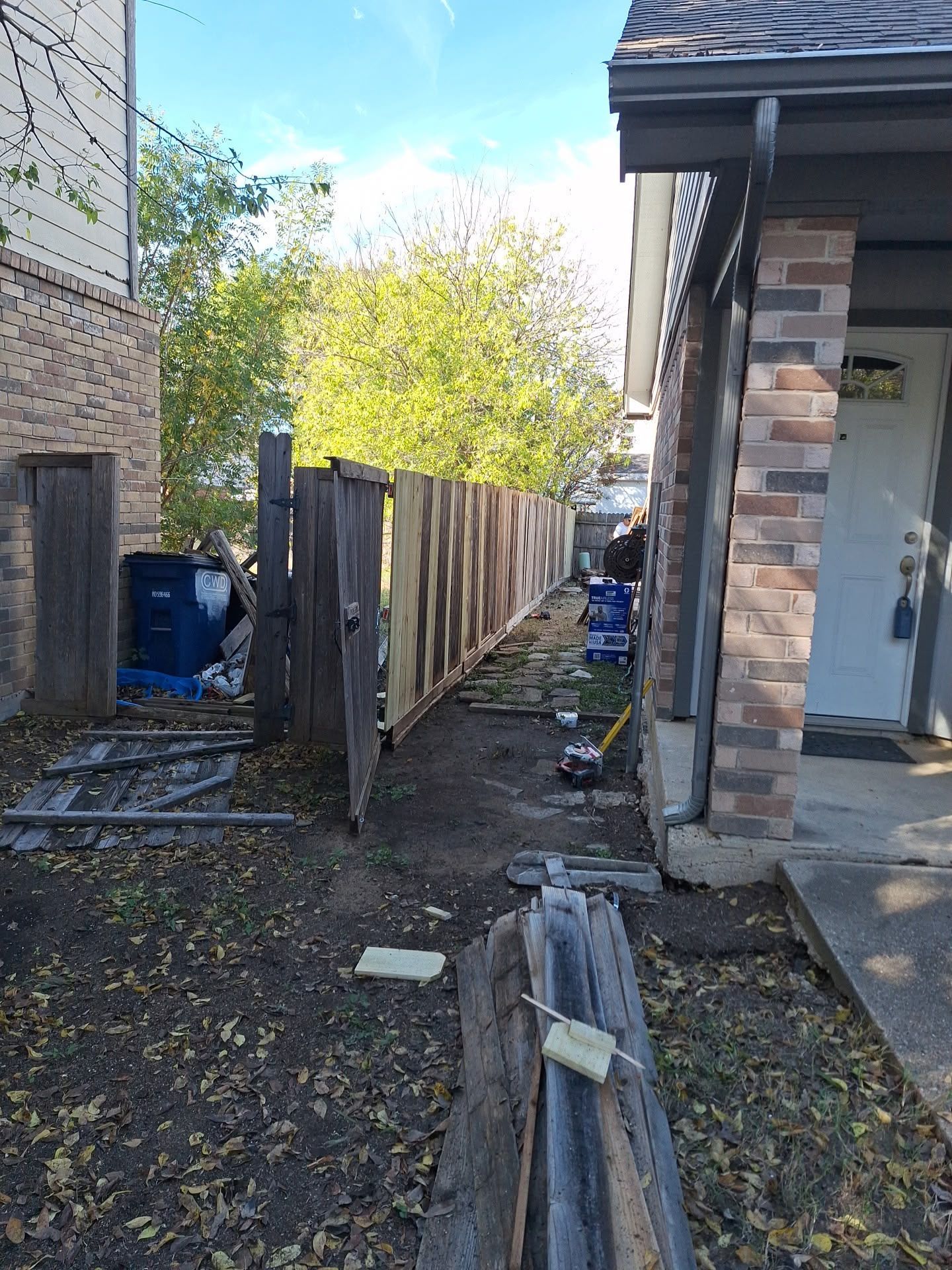 Narrow side yard with a wooden fence, brick house, and scattered debris along a gravel path.