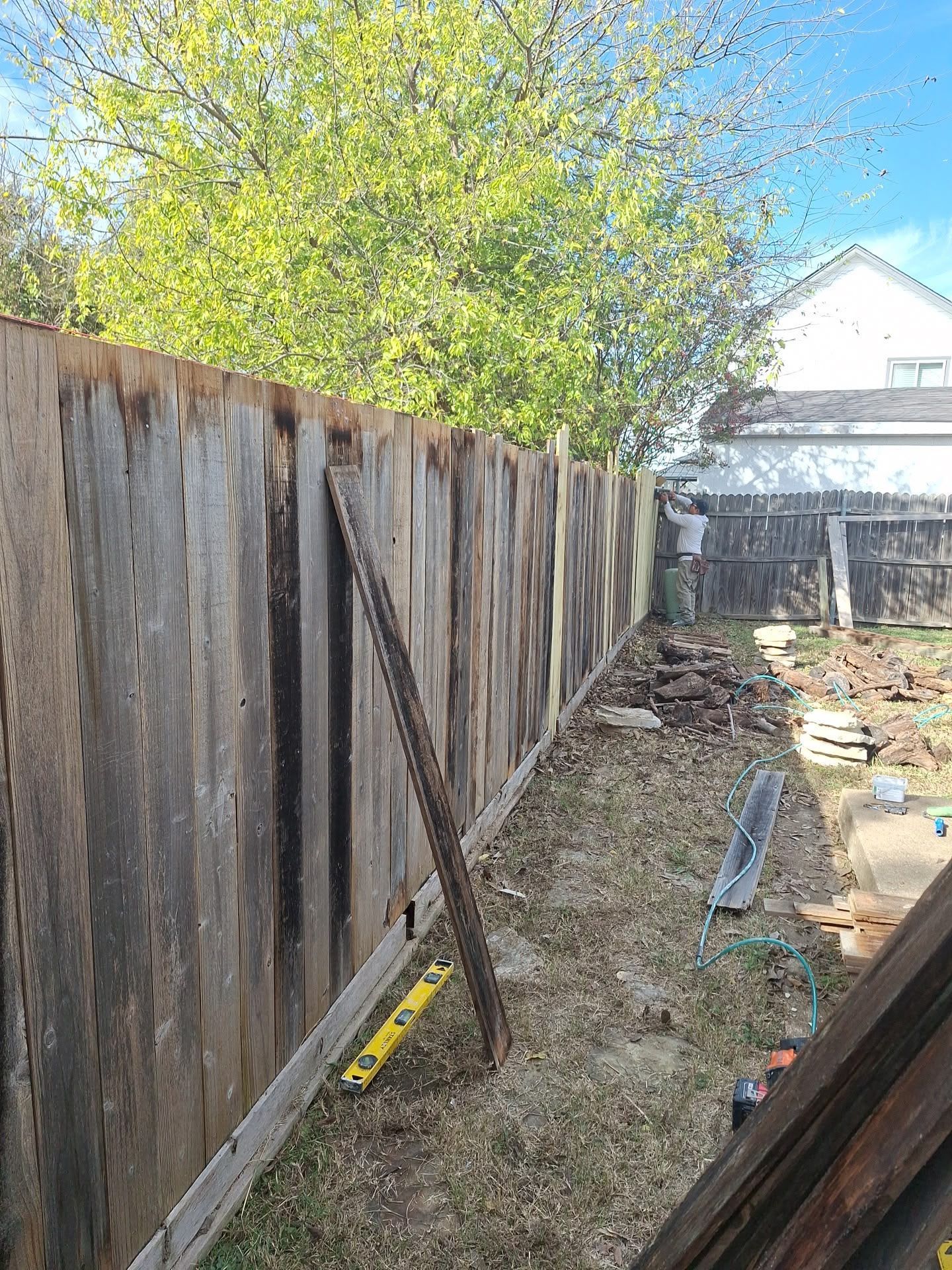 Narrow backyard along a weathered wooden fence with tools and debris on the ground under a bright blue sky