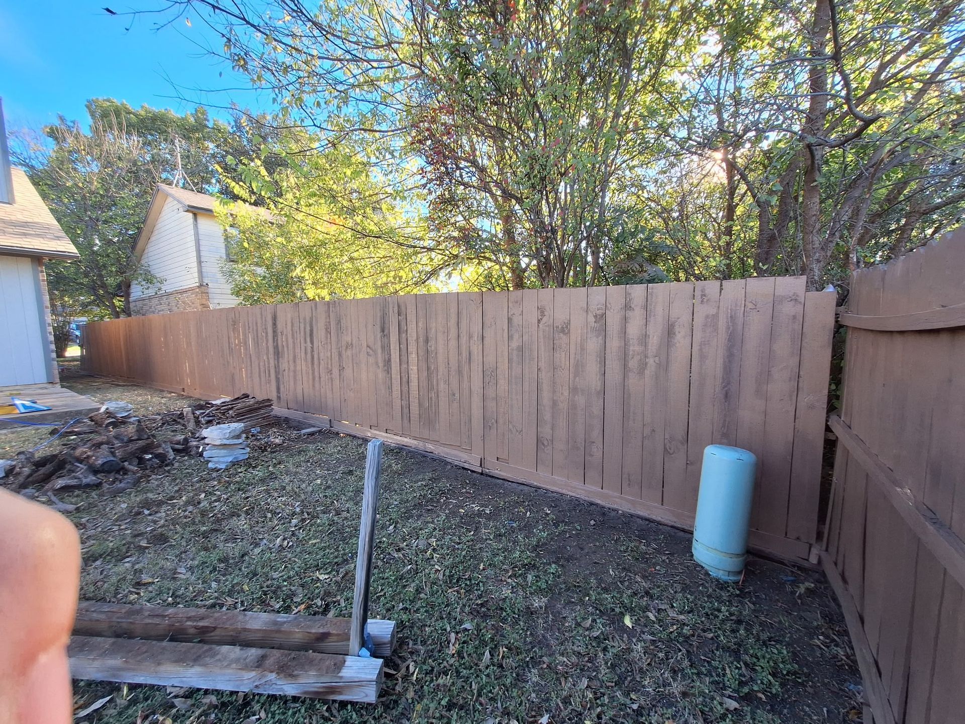 Backyard with tall wooden fence, leaf-covered ground, and a white pipe leaning near the fence.
