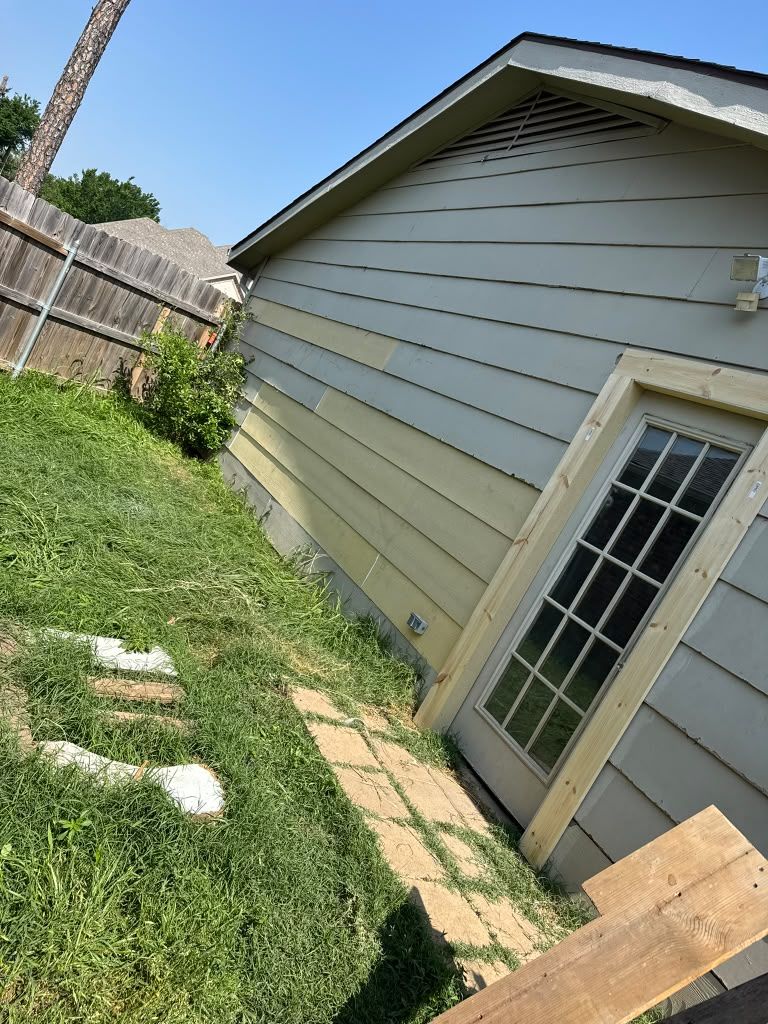 Side of a gray house with a French door, grassy yard, stepping stones, and a wooden board in the foreground