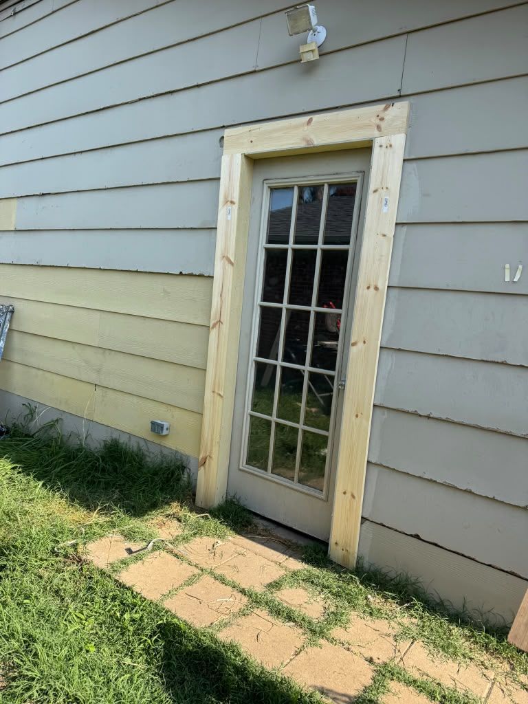 Side of a house with a narrow glass door, tan and gray siding, and a small brick path.