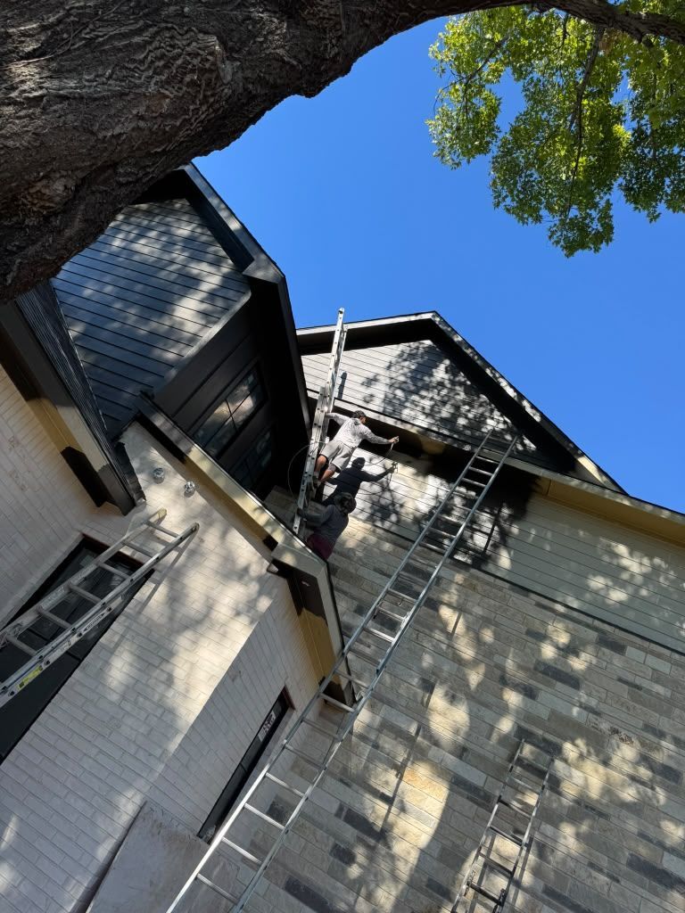 Upward view of white house walls and dark roof against a blue sky, framed by tree branches and shadows