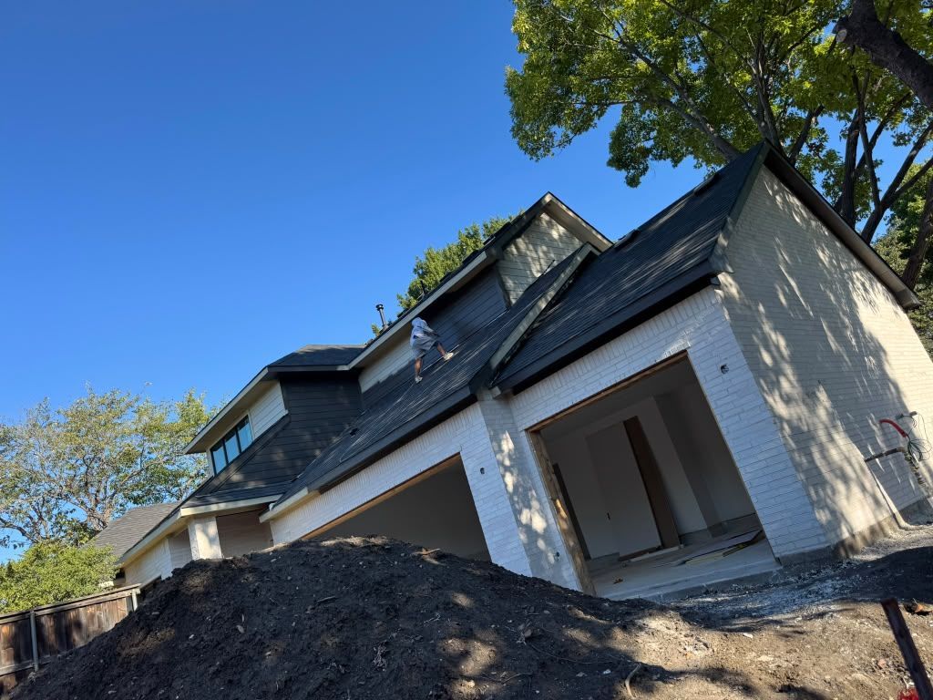 White house under construction with dark roof, large dirt pile, and trees against a blue sky