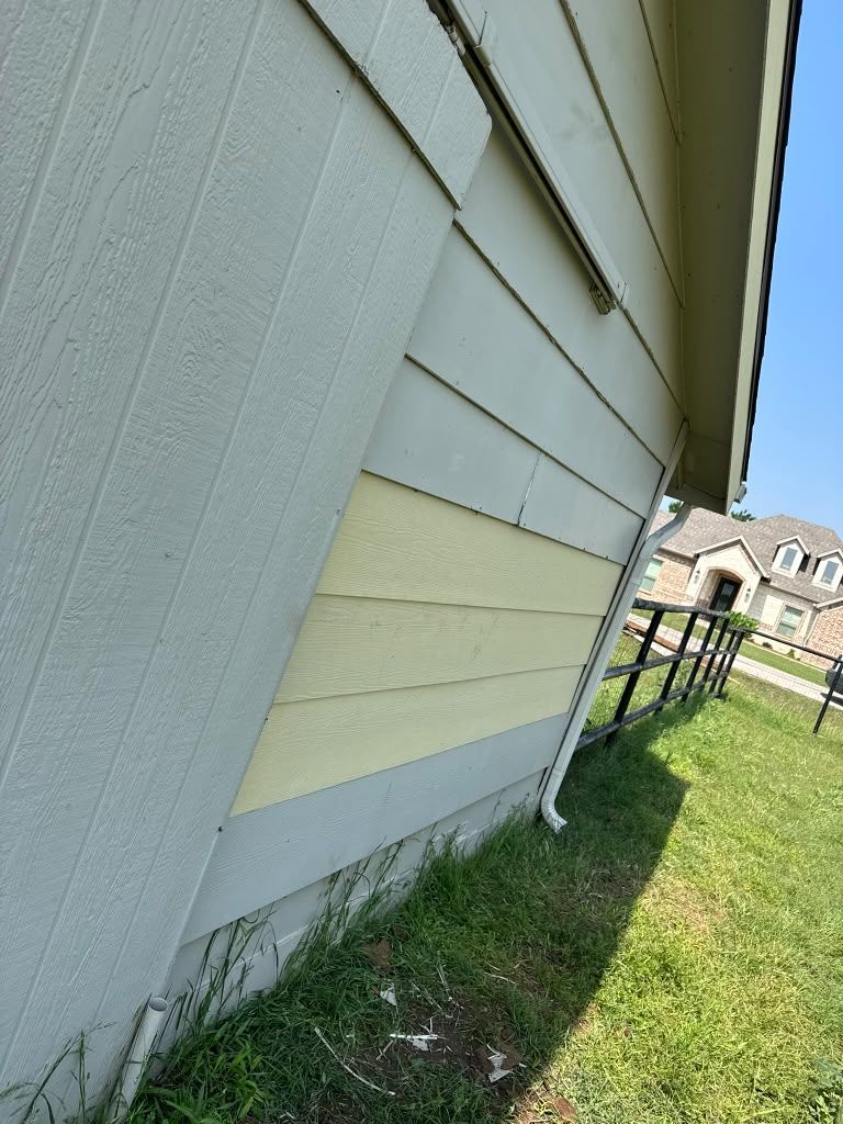 Side of a white house with gray siding, a yellow repair panel, and grass beside a fence under blue sky.