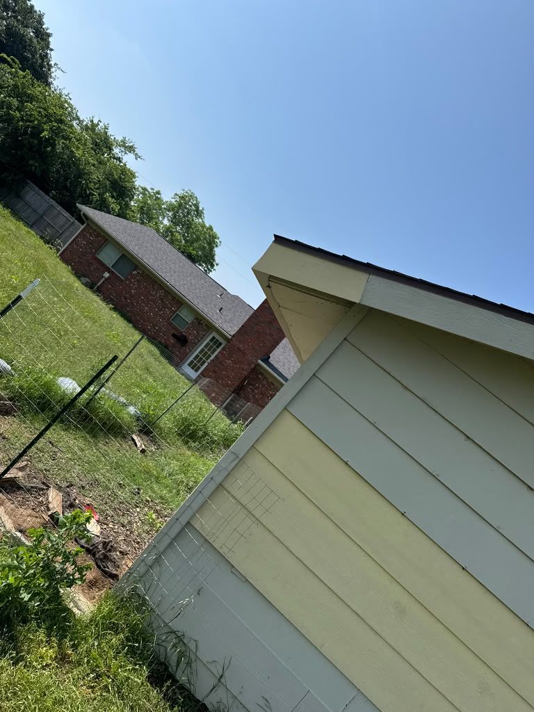 Tilted view of a backyard with a pale yellow shed, fence, grass, and brick houses under a blue sky