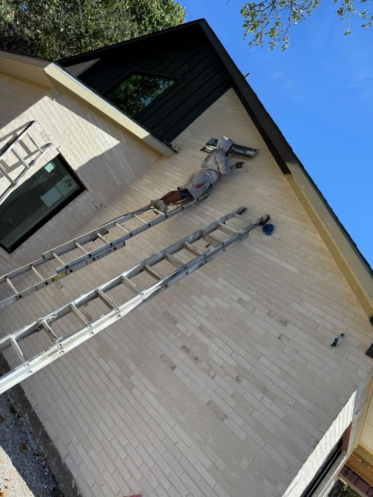 Worker on two extension ladders painting a tan brick building wall near the roofline.