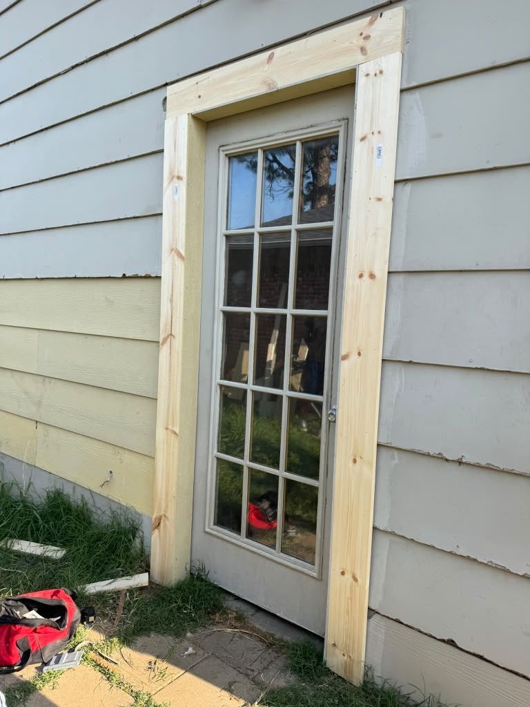 Glass-paneled door set in a light blue house wall with unfinished wood trim and a red tool on the ground.