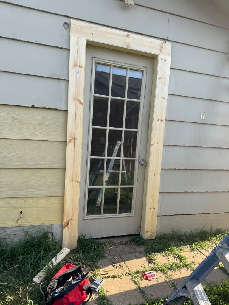 Back door with glass panes in weathered siding, beside a sidewalk and a red-and-black bag.