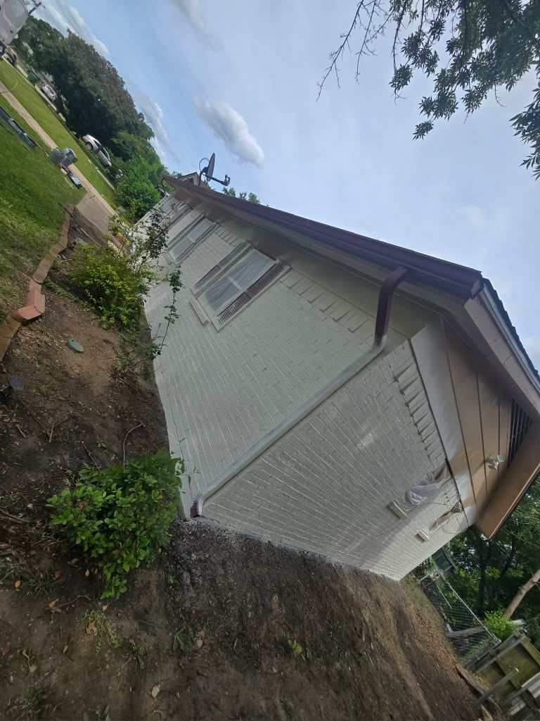 Tilted view of a small white house with a brown roof beside a dirt yard and greenery