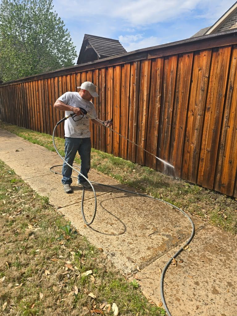 Person pressure-washes a wooden fence beside a sidewalk on a sunny day.