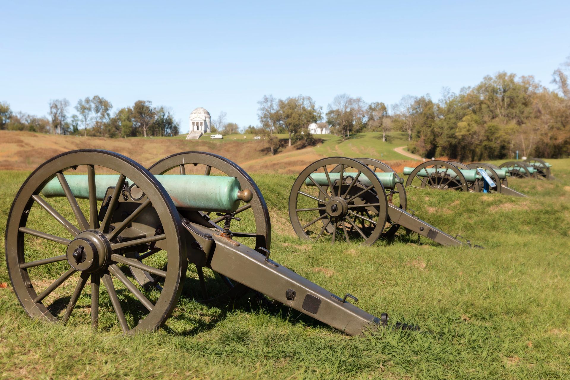 Civil War cannons lined up on a grassy field near a white monument under a clear sky