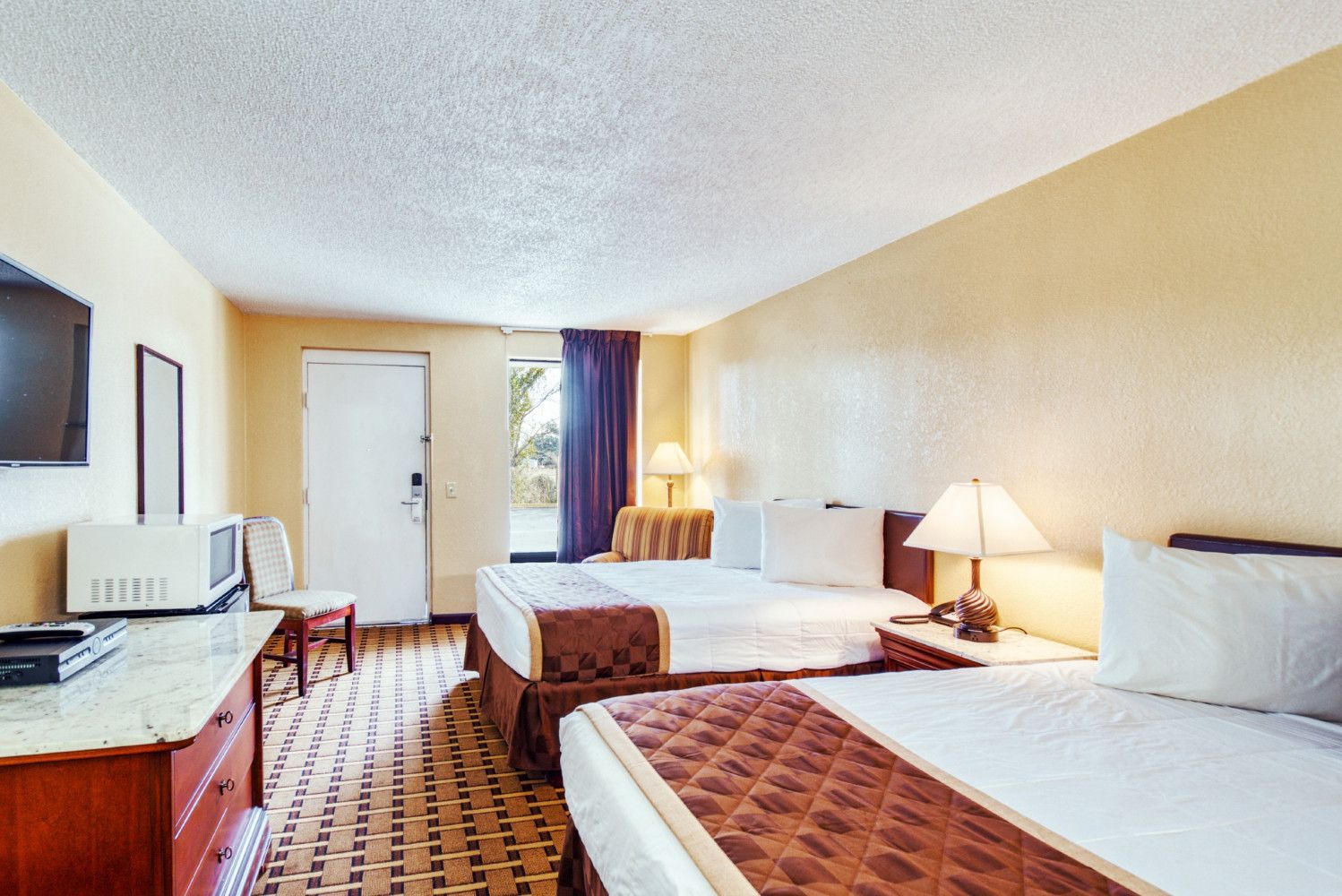 Motel room with two beds, desk, lamp, and window with curtains in warm lighting