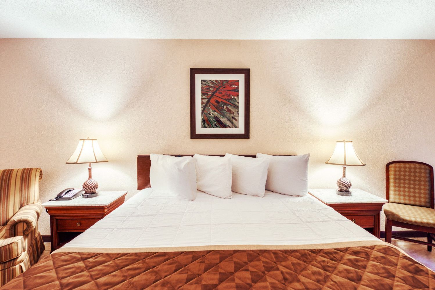Neatly made bed in a warm, symmetrical hotel room with bedside lamps and framed wall art.