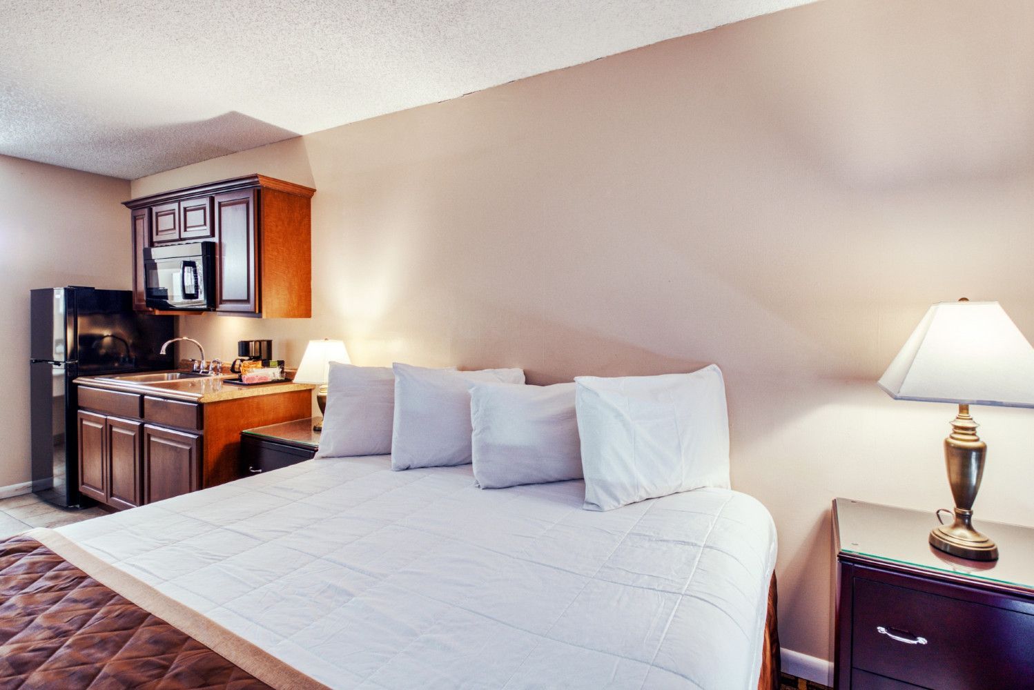 Hotel room with a large white bed, dresser, lamp, and compact kitchenette.