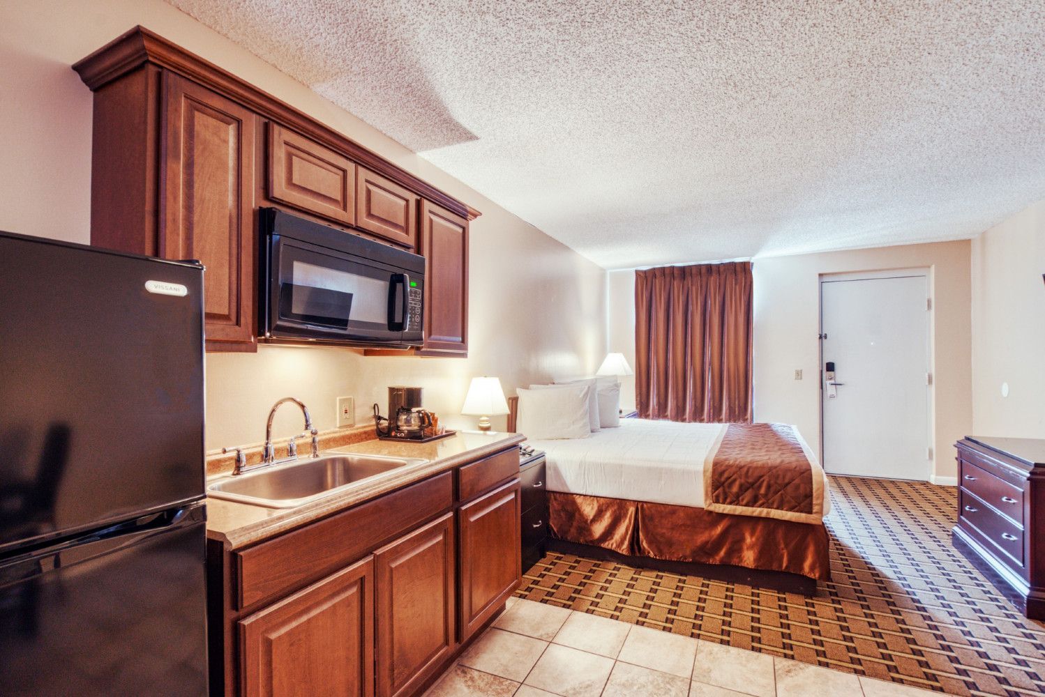 Small hotel room with kitchenette, bed, dresser, and window curtains