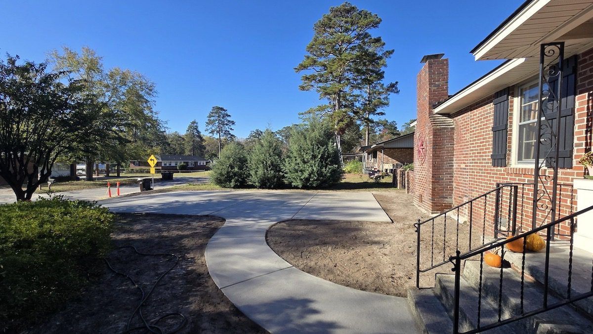 A concrete walkway leads to the side entrance of a red brick house with a chimney, set against a yard with mature trees.