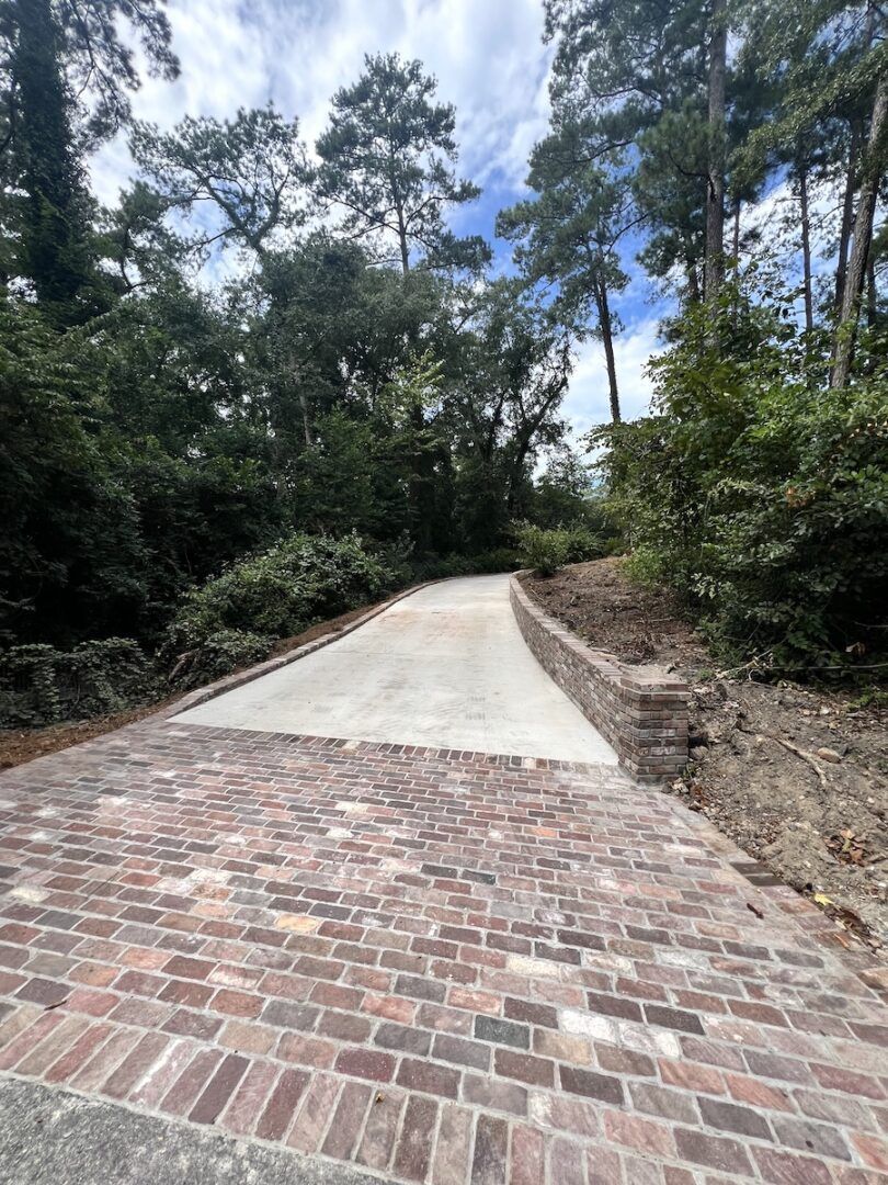 A paved driveway transitioning from red brick pavers to smooth concrete, winding uphill through a wooded landscape.