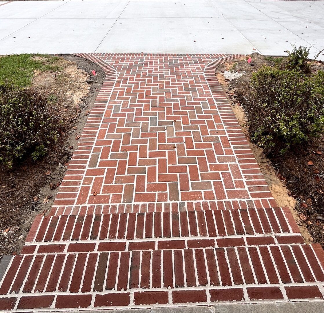 A brick walkway with a herringbone pattern leads to a concrete driveway, flanked by green bushes on both sides.