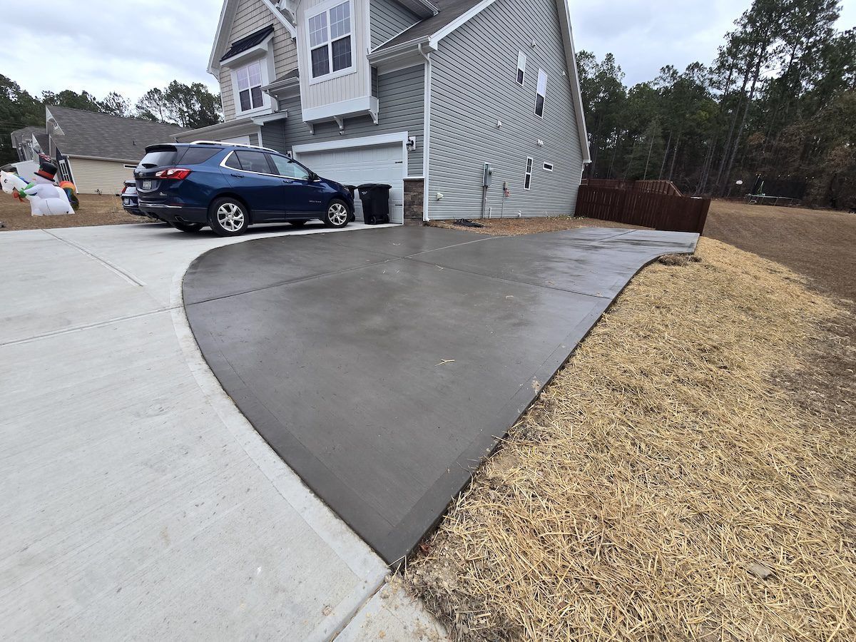 A newly poured, dark gray concrete driveway extension sits next to a lighter gray existing driveway by a house.