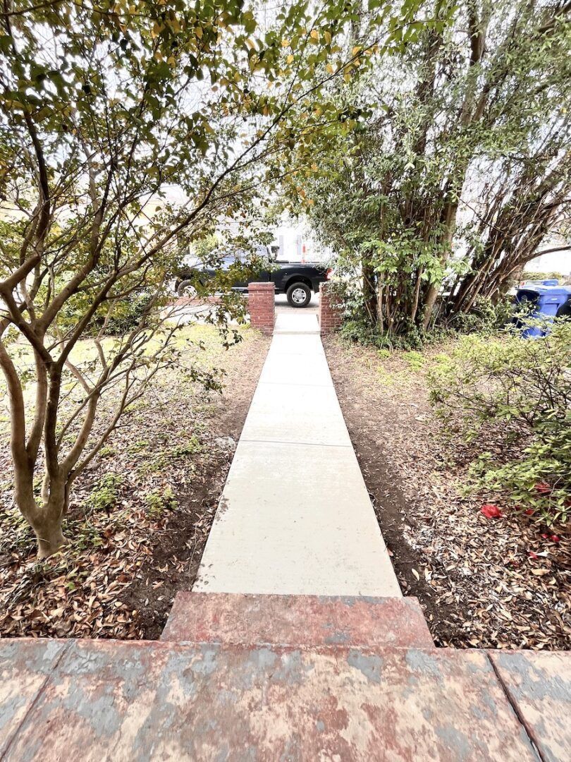A concrete walkway leads from the foreground through trees and bushes toward a parked car in the distance.