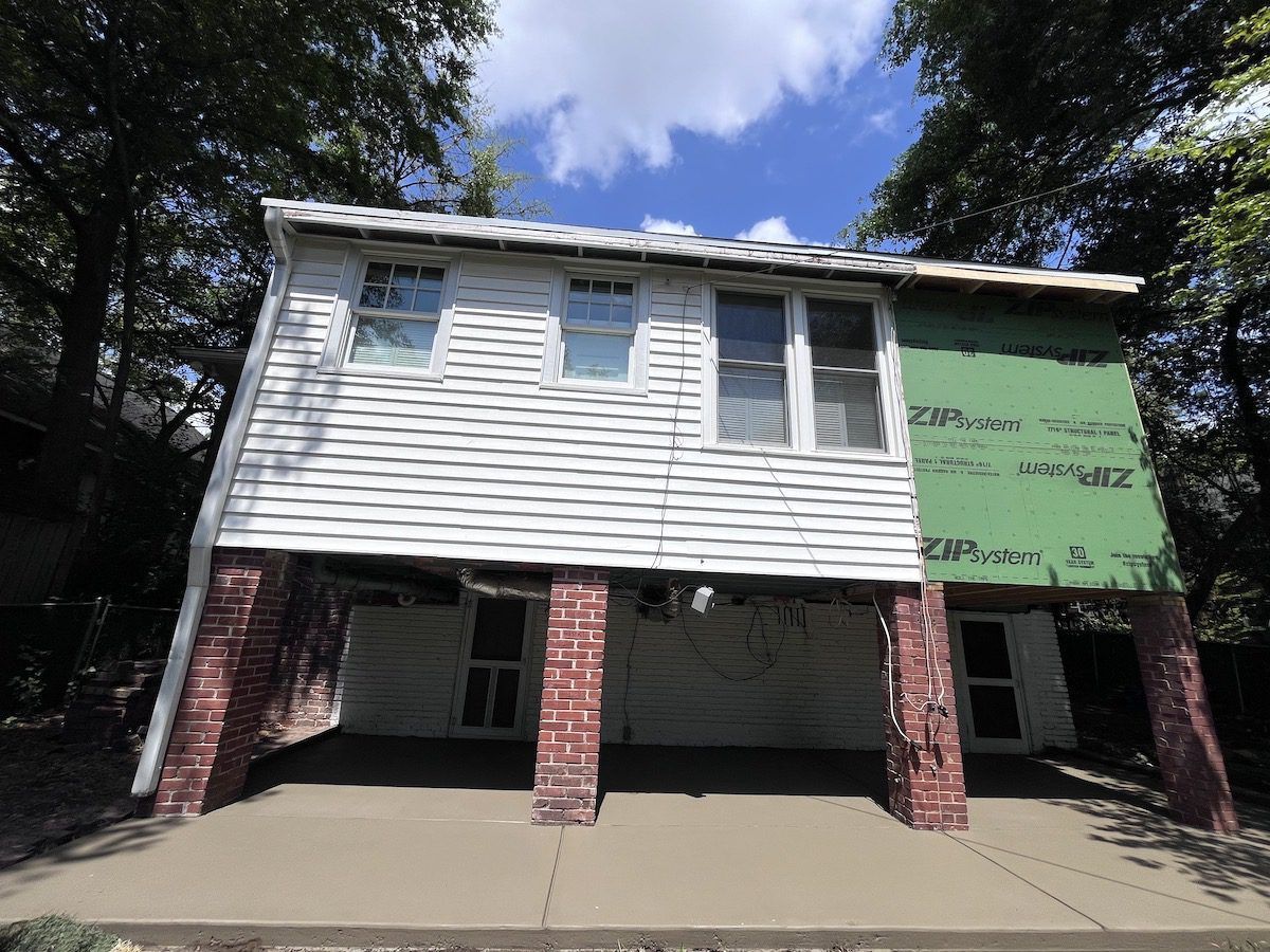 A two-story house exterior under construction, with white siding, unfinished green panels, and a brick-pillared base.