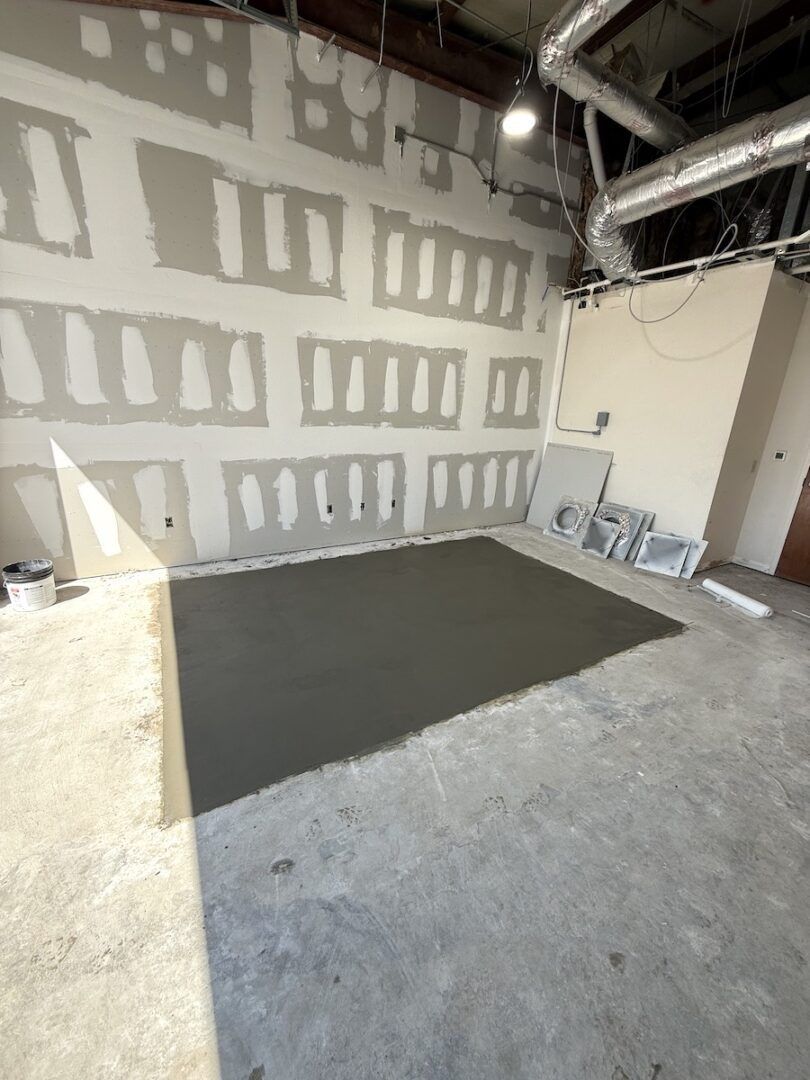A room under renovation with fresh concrete flooring, drywall with tape, and exposed ceiling ductwork.