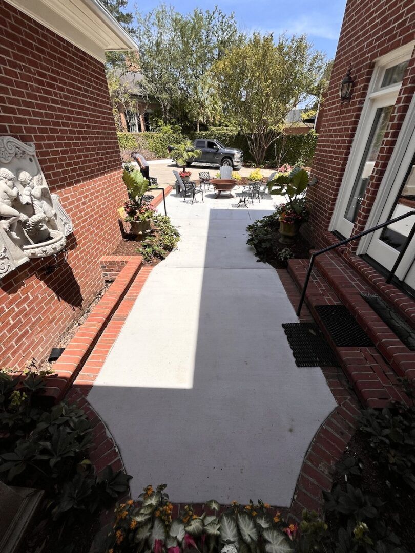 A narrow, light-colored concrete walkway between two brick walls leads to a sunny outdoor patio with chairs and plants.