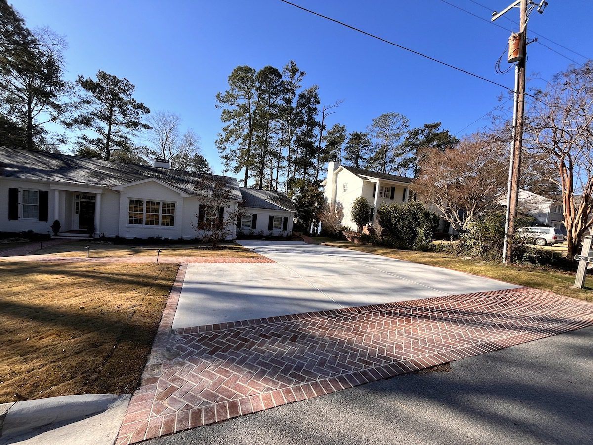 A concrete driveway with a brick border leads to a white house under a clear blue sky, surrounded by trees.