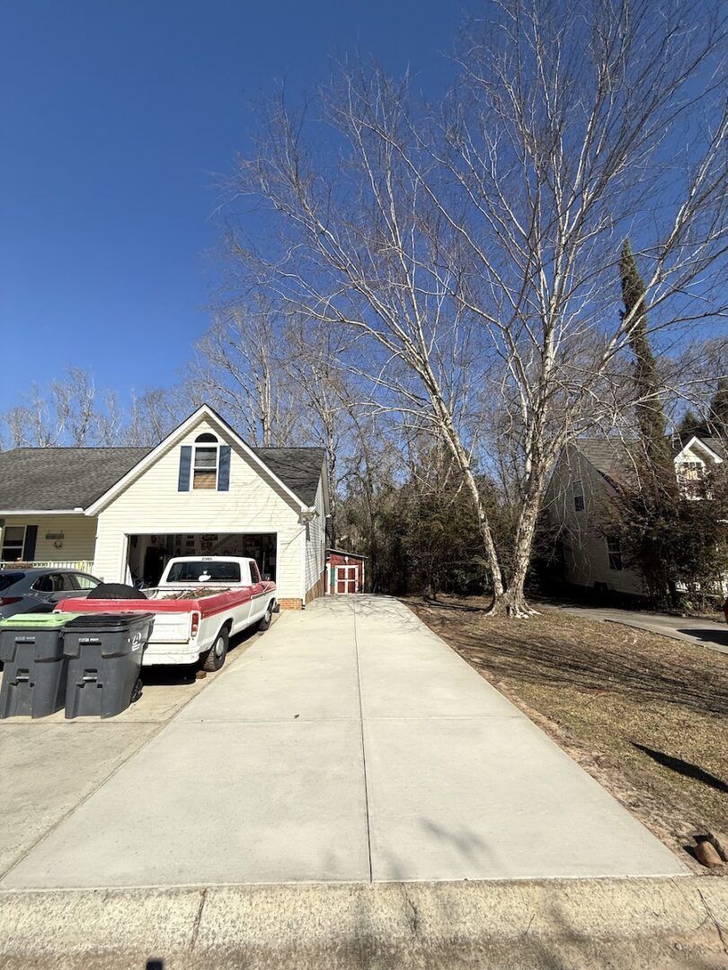 A concrete driveway leads to a garage with a parked white pickup truck and two trash bins; trees frame the background.