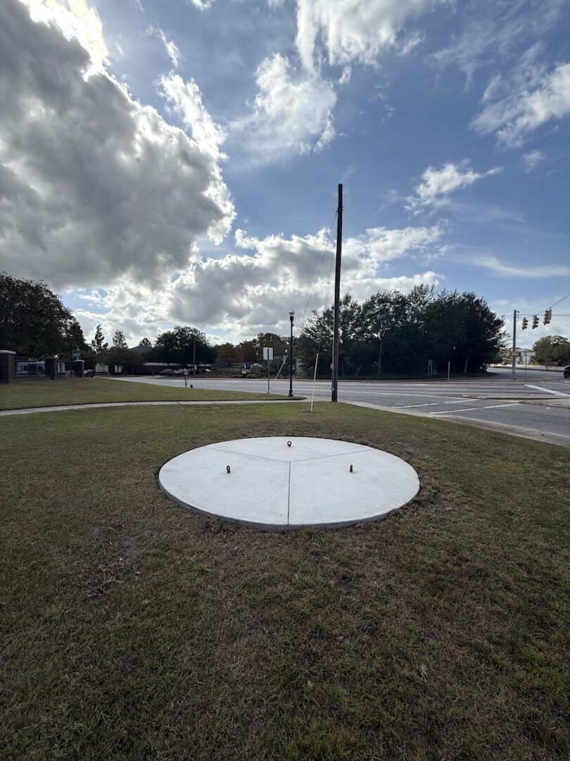 A flat, white, circular concrete pad set in a grassy roadside area under a partly cloudy sky.