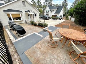 A residential patio area with a white house, a woven patio table and chairs, and a covered grill on a stone surface.