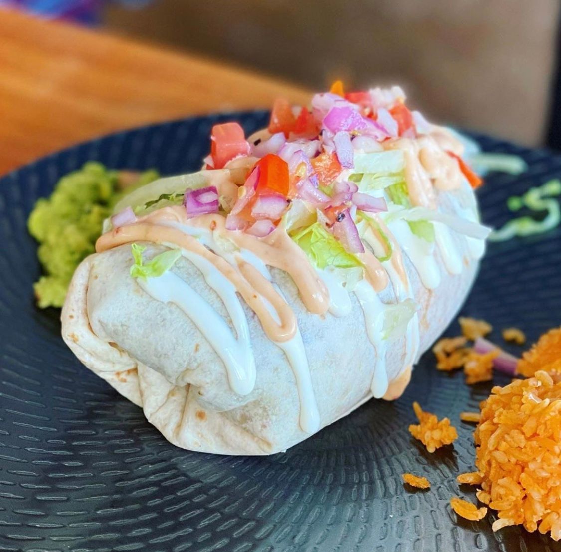 Burrito with toppings, guacamole, and rice on a black plate.