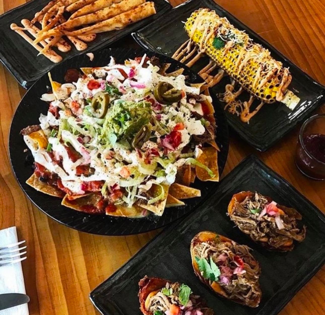 A spread of Mexican food on black plates: nachos, corn on the cob, egg rolls, and fries on a wooden table.