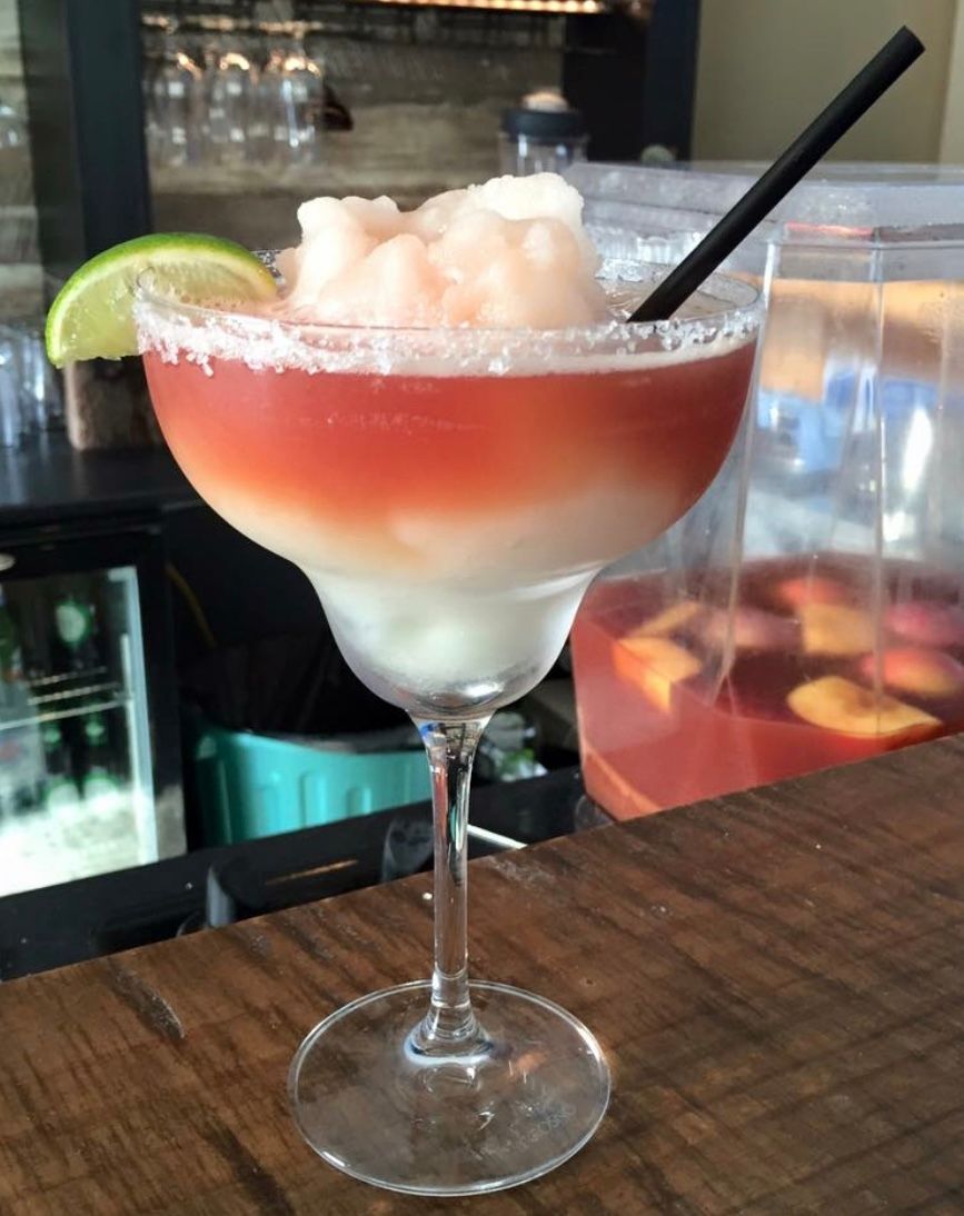 Cocktail in a margarita glass with pink and white layers, garnished with lime and straw, on a wooden bar.