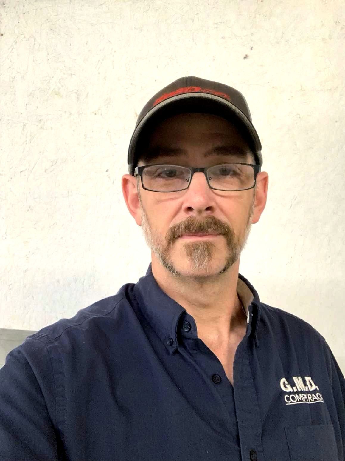 A man wearing glasses and a hat is standing in front of a white wall.