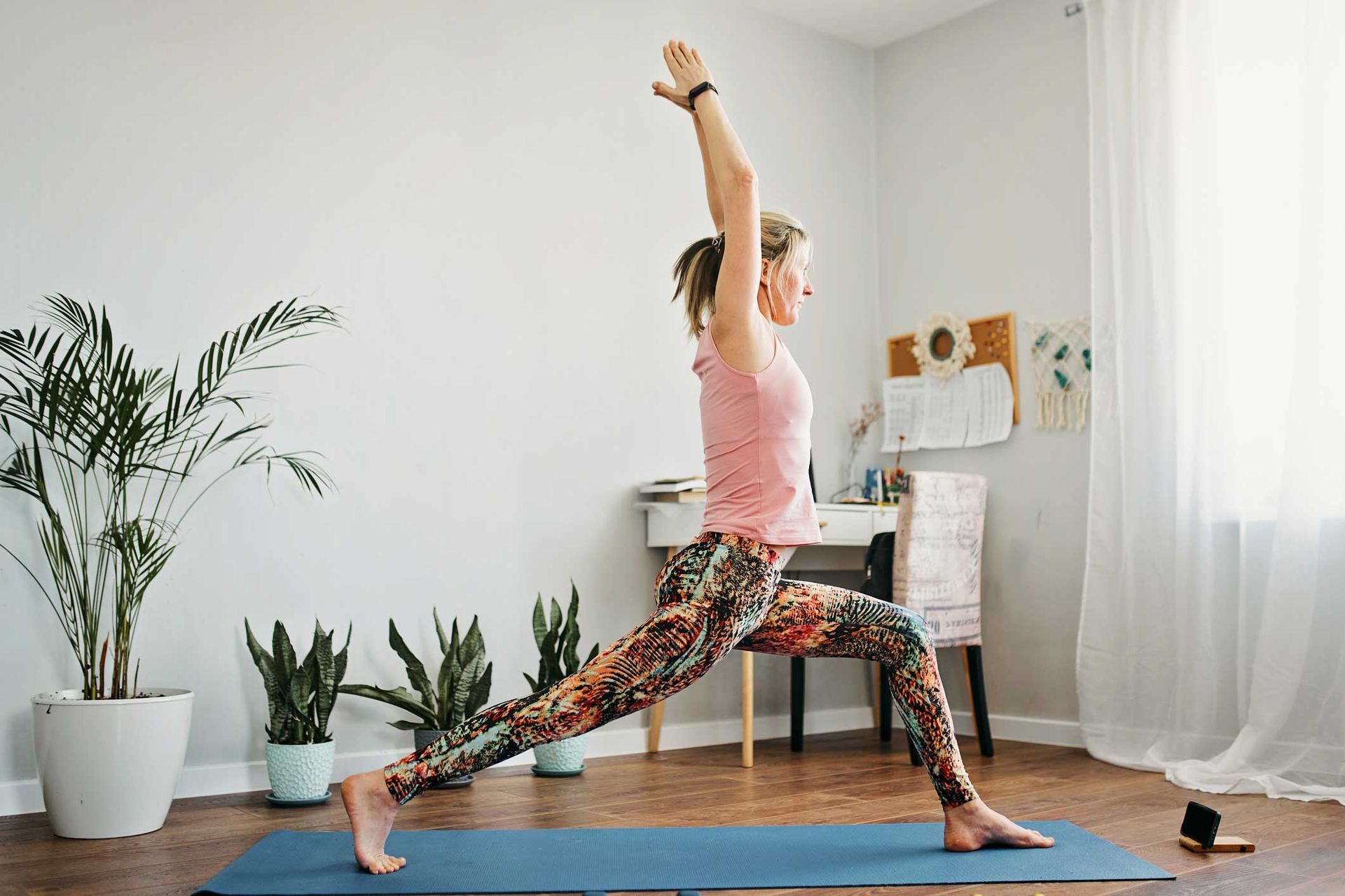Woman in yoga pose, arms raised, in a brightly lit room with plants and a desk. Woman in yoga pose, arms raised, in a brightly lit room with plants and a desk.