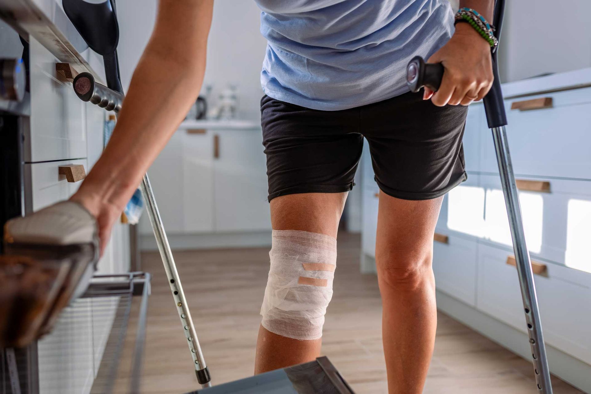 Person with bandaged knee uses crutches and holds onto furniture for support in a kitchen. Person with bandaged knee uses crutches and holds onto furniture for support in a kitchen.